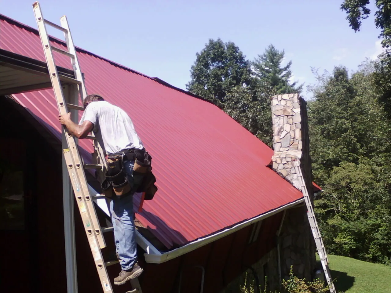 Roofer on a ladder beside a red metal roof and stone chimney — Soffit & Fascia Repair by Roofing Brothers in West Chazy
